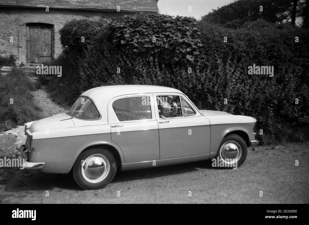 1950s, historical, on a rural lane, a little boy sitting in the driver's seat of a family car, a Hillman Minx, waving, England, UK. Founded in 1907, the Hillman Motor car company was based near Coventry and the Hillman Minx was in production from 1931 to 1970, with many different versions. The car seen here is possibly the Minx series V. Hillman, originally a bicycle maker, was taken over by the Rootes Brothers in 1928. Stock Photo