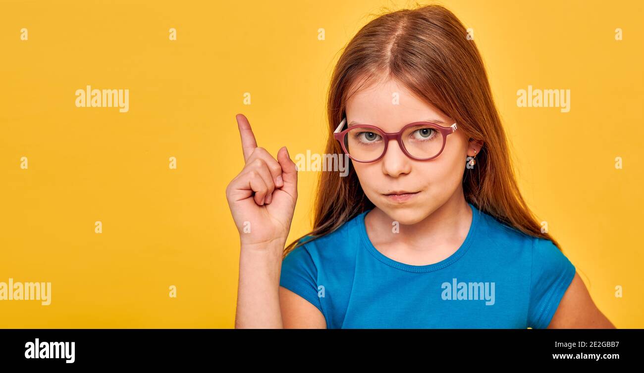 Cute girl wearing eyeglasses and blue t-shirt, with serious emotions on her face, pointing finger into empty space on yellow background Stock Photo