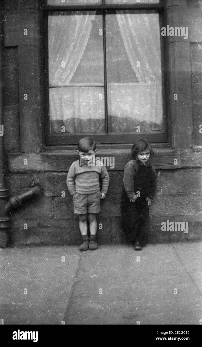 1940s, historical, two small children, perhaps a brother and little sister, standing on the pavement outside a house below a large victorian wooden sash window with net curtain, England, UK. Stock Photo