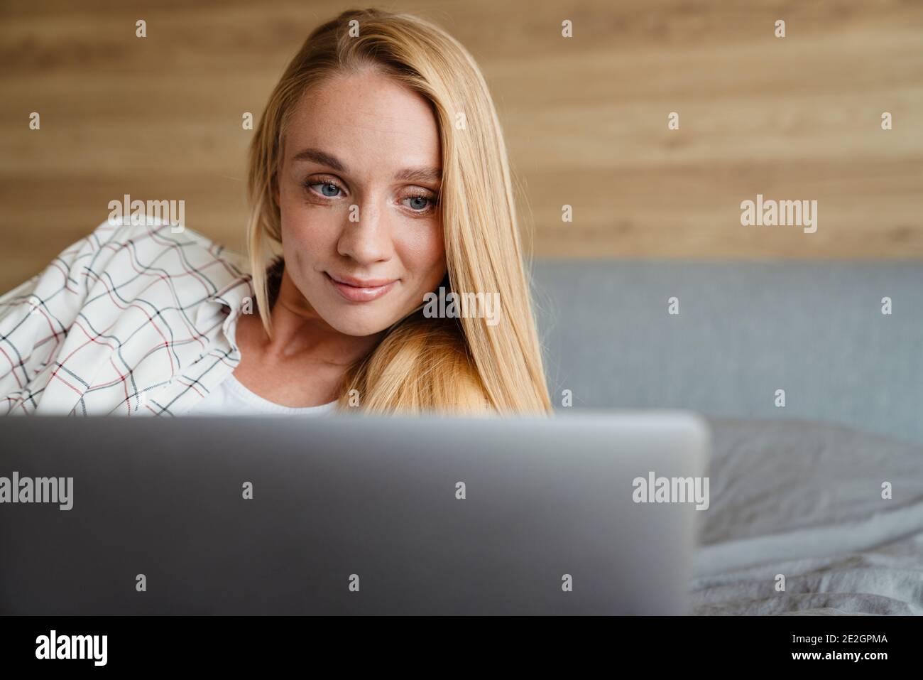 Beautiful blonde smiling woman using laptop while lying on bed at home ...