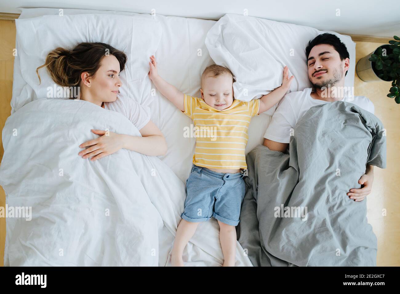 Toddler stretching out his arms, lying between his parents in bed Stock