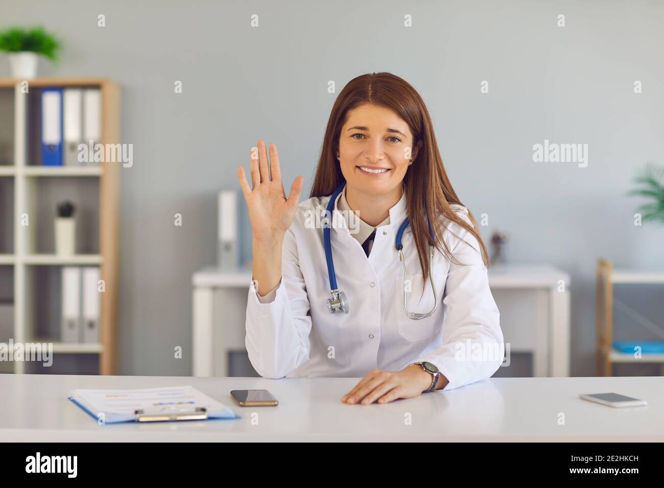 Female doctor waving hand greeting his viewers starting an online broadcast in a hospital office. Stock Photo