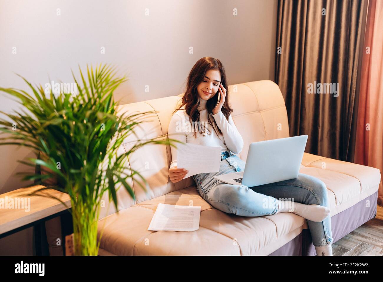 Freelance student young female using laptop while sitting at home. Young woman sitting on sofa and working online on laptop. Pretty female talking on Stock Photo