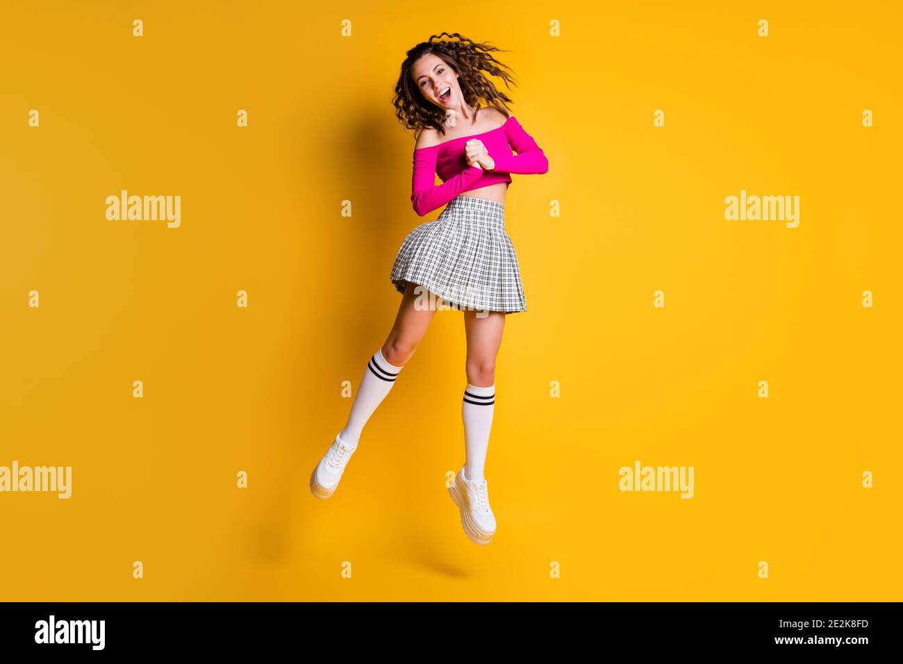 Photo portrait of curly brunette girl jumping holding hands together screaming wearing casual fuchsia crop-top plaid skirt knee-high socks white shoes Stock Photo