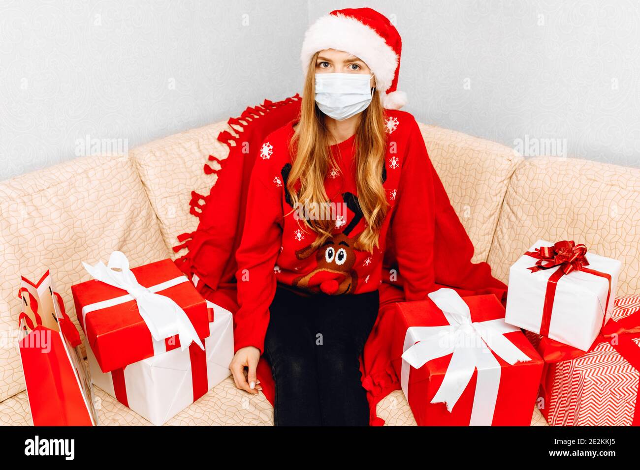 Young woman in medical mask and Santa Claus hat, greets Christmas New Year at home while sitting on the sofa with Christmas gifts Stock Photo