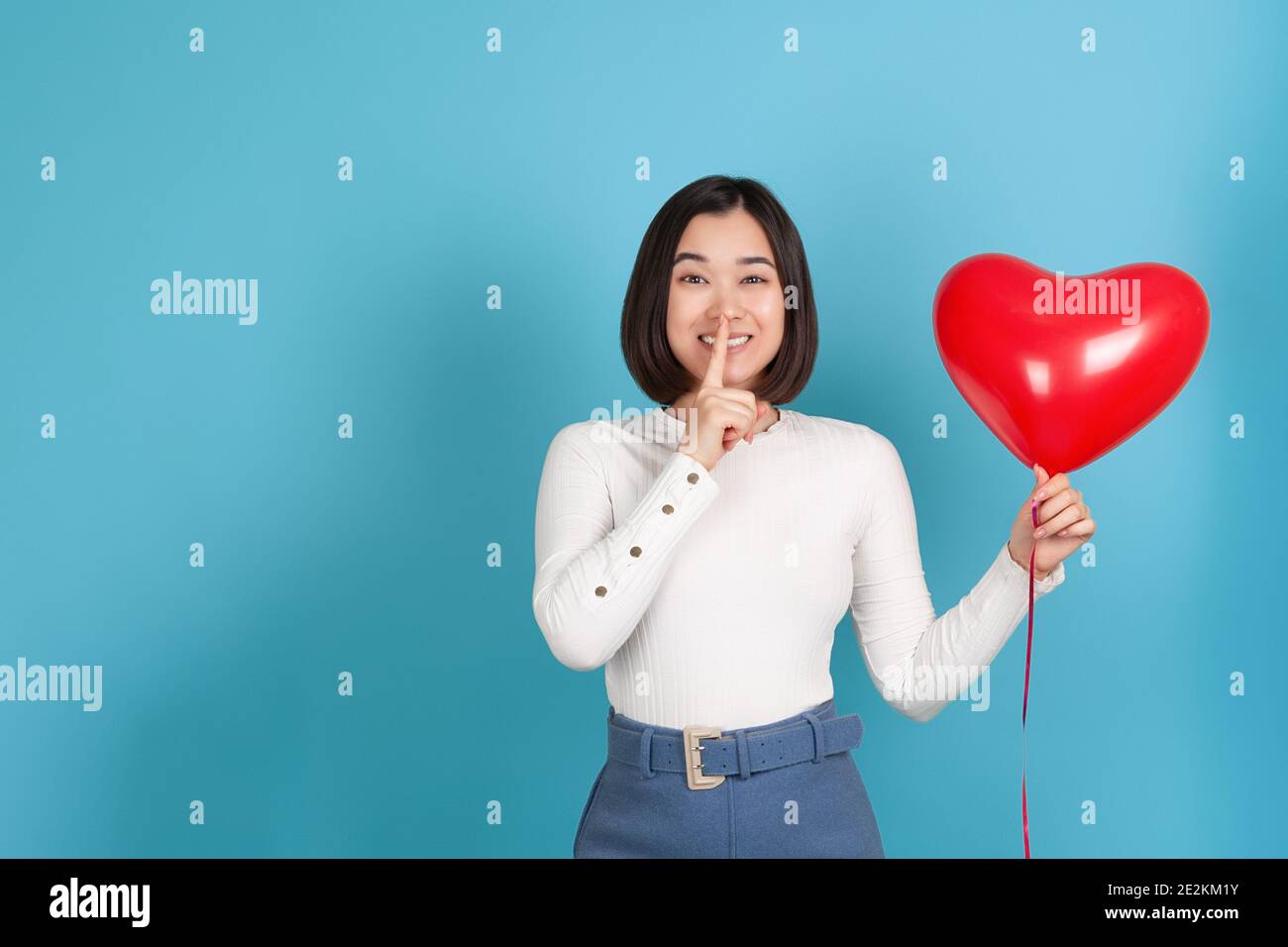 Naughty, playful young Asian woman holds a flying heart-shaped balloon and index finger near her mouth, isolated on a blue background Stock Photo