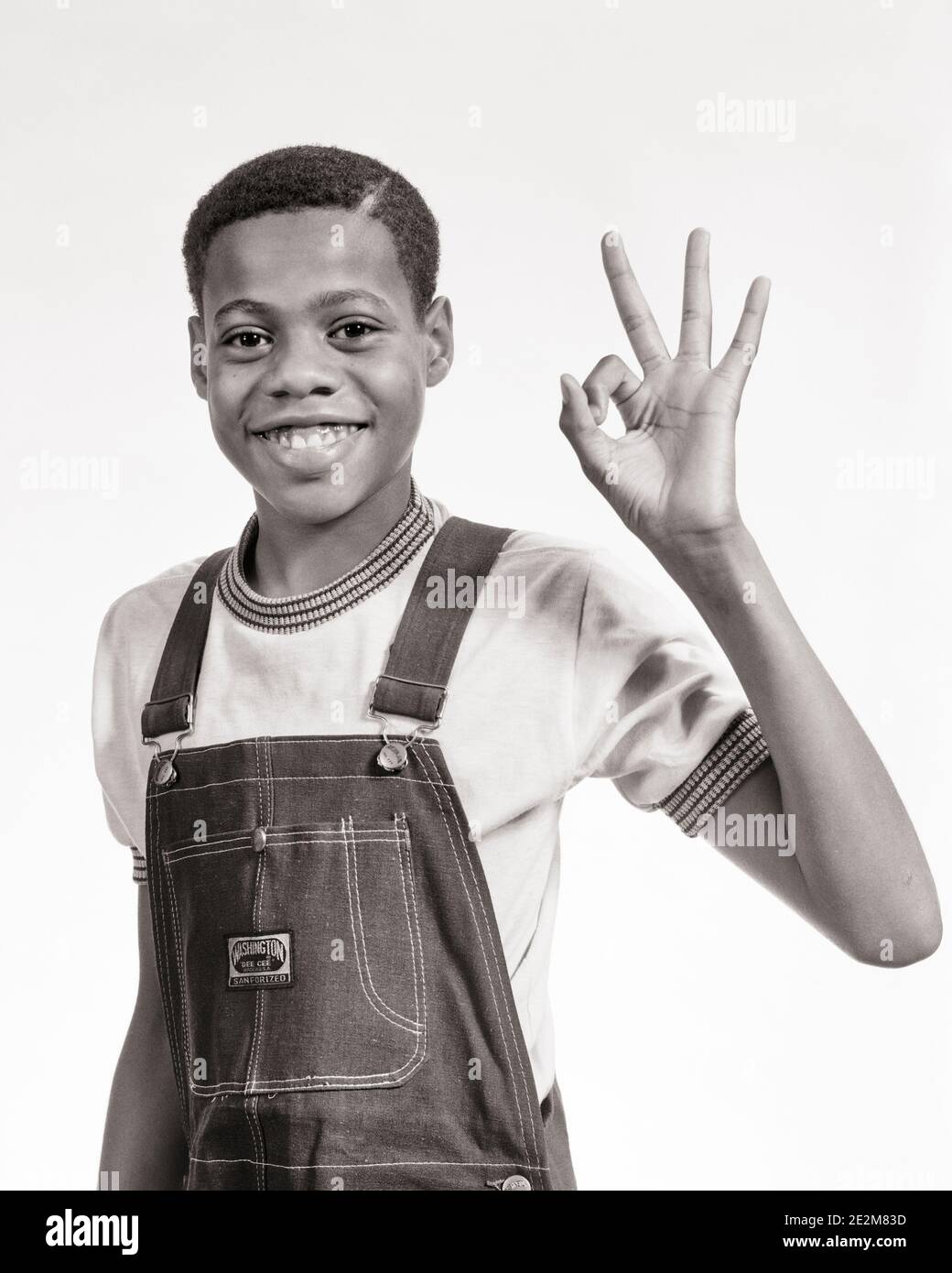 1970s SMILING AFRICAN-AMERICAN PRE-TEEN BOY WEARING BIB OVERALLS MAKING OKAY SIGN WITH HAND LOOKING AT CAMERA - b25650 HAR001 HARS MALES OK TEENAGE BOY DENIM GESTURING B&W EYE CONTACT DREAMS HAPPINESS CHEERFUL AFRICAN-AMERICANS AFRICAN-AMERICAN OKAY BLACK ETHNICITY GESTURES SMILES CONCEPTUAL JOYFUL BIB FINE INFORMAL JUVENILES OKEY-DOKEY PRE-TEEN PRE-TEEN BOY TWILL BLACK AND WHITE CASUAL HAR001 OLD FASHIONED AFRICAN AMERICANS Stock Photo