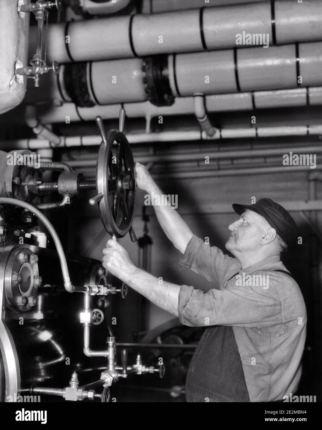 1930s MATURE MAN WORKER REGULATING STEAM PRESSURE OR FLOW BY TURNING THE WHEEL-LIKE HANDLE ON A STEAM VALVE - i1913 HAR001 HARS EMPLOYMENT OCCUPATIONS HANDLE EMPLOYEE OR FLOW BLACK AND WHITE CAUCASIAN ETHNICITY HAR001 LABORING OLD FASHIONED VALVE Stock Photo