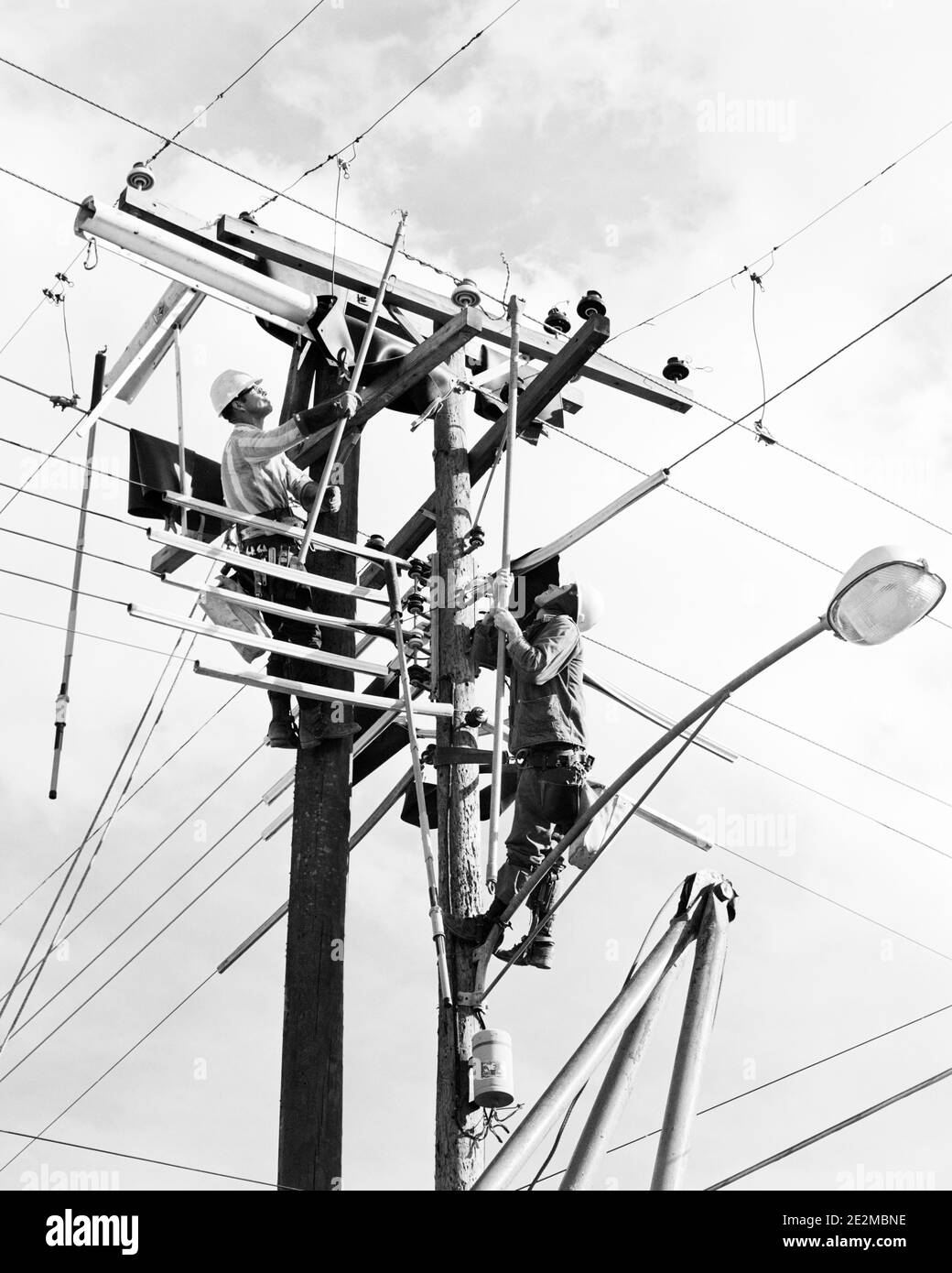 1960s TWO MEN WORKING ON PUBLIC UTILITY ELECTRICAL WIRES AT TOP OF POWER POLES - i5376 HAR001 HARS PROTECTION LABOR EMPLOYMENT LINES OCCUPATIONS MAINTENANCE UTILITY EMPLOYEE LINEMAN STREET LAMP LINEMEN MID-ADULT MID-ADULT MAN YOUNG ADULT MAN BLACK AND WHITE CAUCASIAN ETHNICITY HAR001 LABORING OLD FASHIONED POWER LINES PUBLIC UTILITY Stock Photo