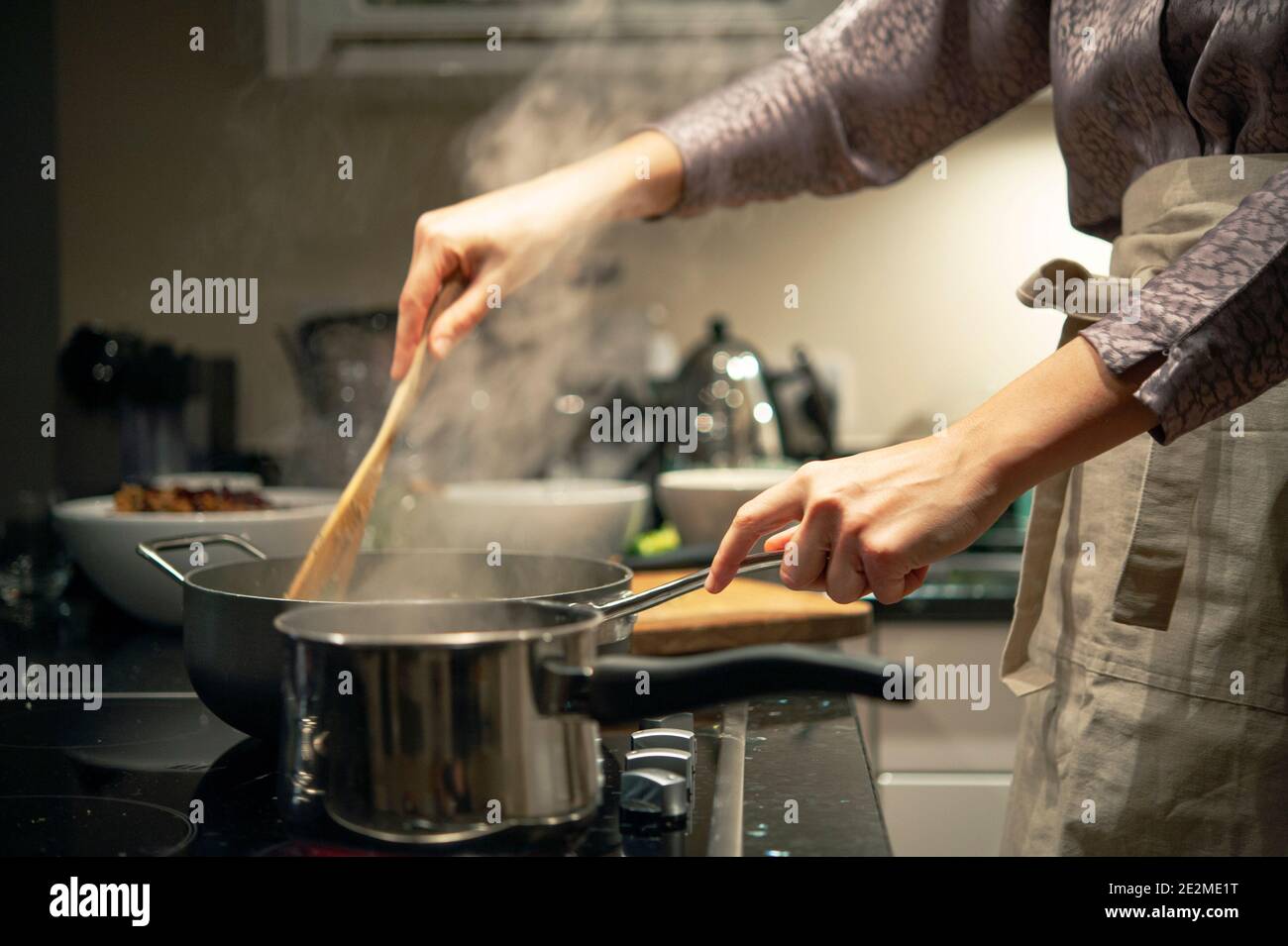 Woman in apron cooking on ceramic hob in modern home, close-up of hands stirring pot in kitchen. London, UK Stock Photo