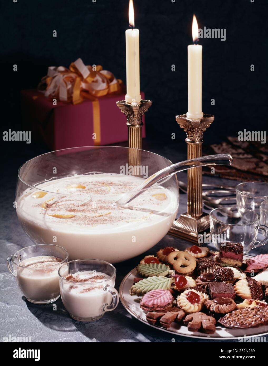 1980s CUPS LADLE AND BOWL OF CHRISTMAS EGGNOG BEVERAGE WITH PLATE OF SUGAR COOKIES LIT CANDLES AND A WRAPPED PRESENT - kx9988 DAS001 HARS DECEMBER DECEMBER 25 STILL LIFE BOURBON RUM PUNCH BOWL WHISKY BEVERAGES FESTIVE JANUARY 1 LIGHTED NEW YEAR NEW YEARS OLD FASHIONED Stock Photo