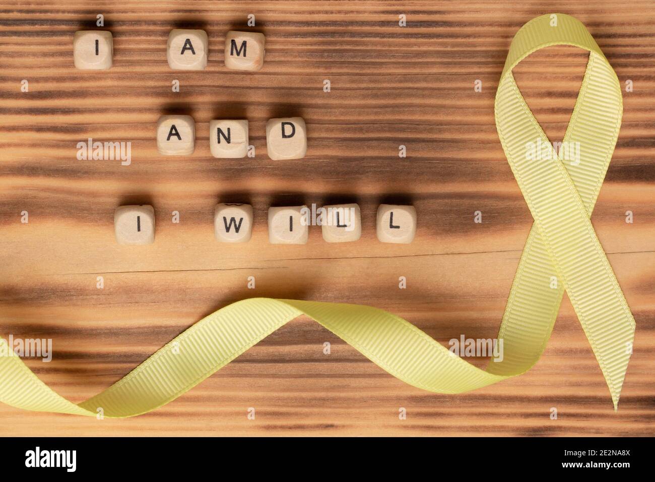 Wooden cubes with text of the slogan of World Cancer Day I am and I will and a yellow ribbon, on a wooden surface. Flat lay Stock Photo
