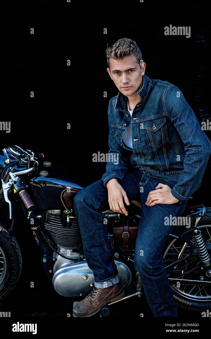 Handsome man wearing double denim sitting on motorcycle with black background . Portrait of handsome young man on vintage cafe racer motorcycle . Stock Photo
