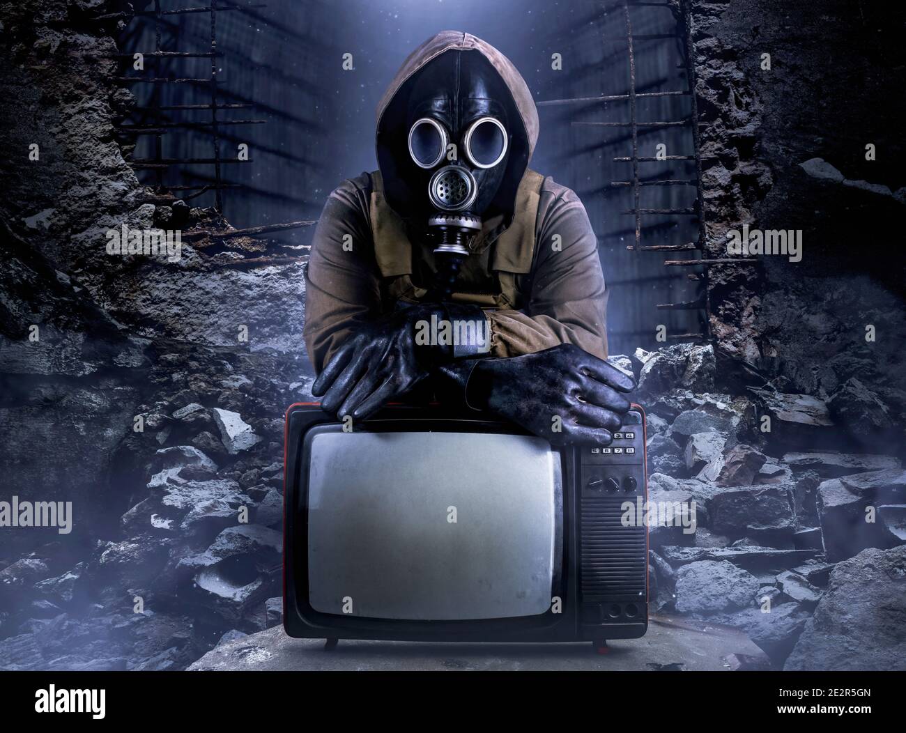 Photo of stalker soldier in jacket and armored vest and rubber gloves standing with old tv set on ruined dark background. Stock Photo