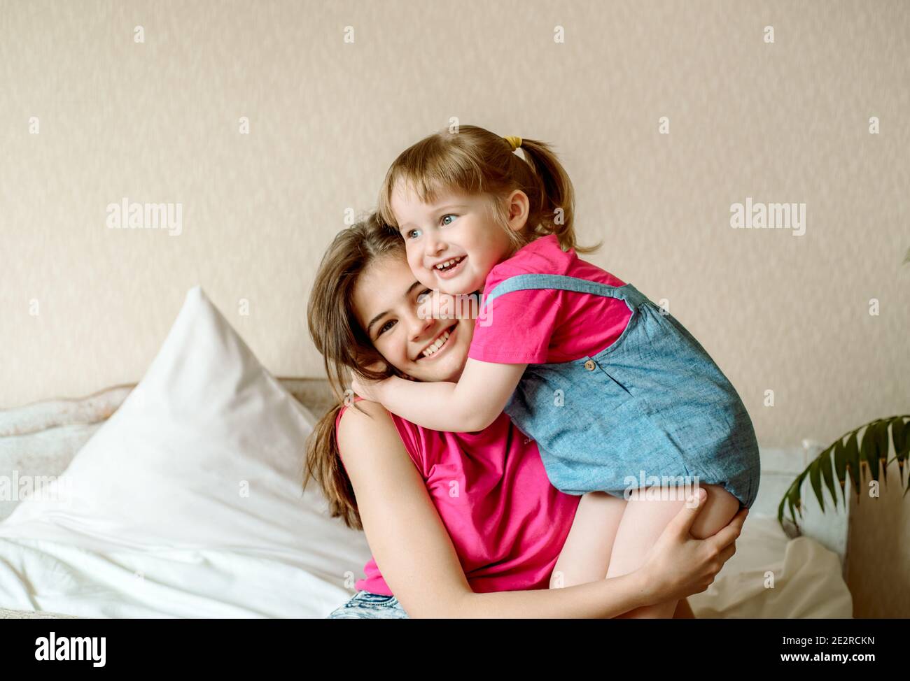 Happy kids having fun with pillows on bed.children play in the bedroom. two sisters. teenager and baby girl laugh, hug and kiss. Stock Photo