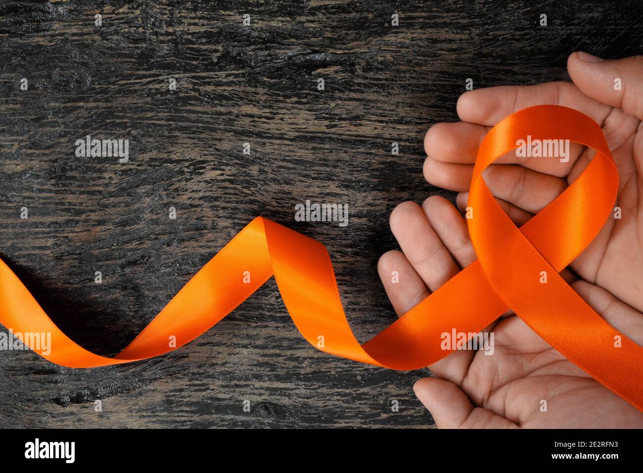Top view of male hand holding orange ribbon on dark wood background. Leukemia, kidney cancer, adhd, copd, malnutrition and hunger awareness. Stock Photo