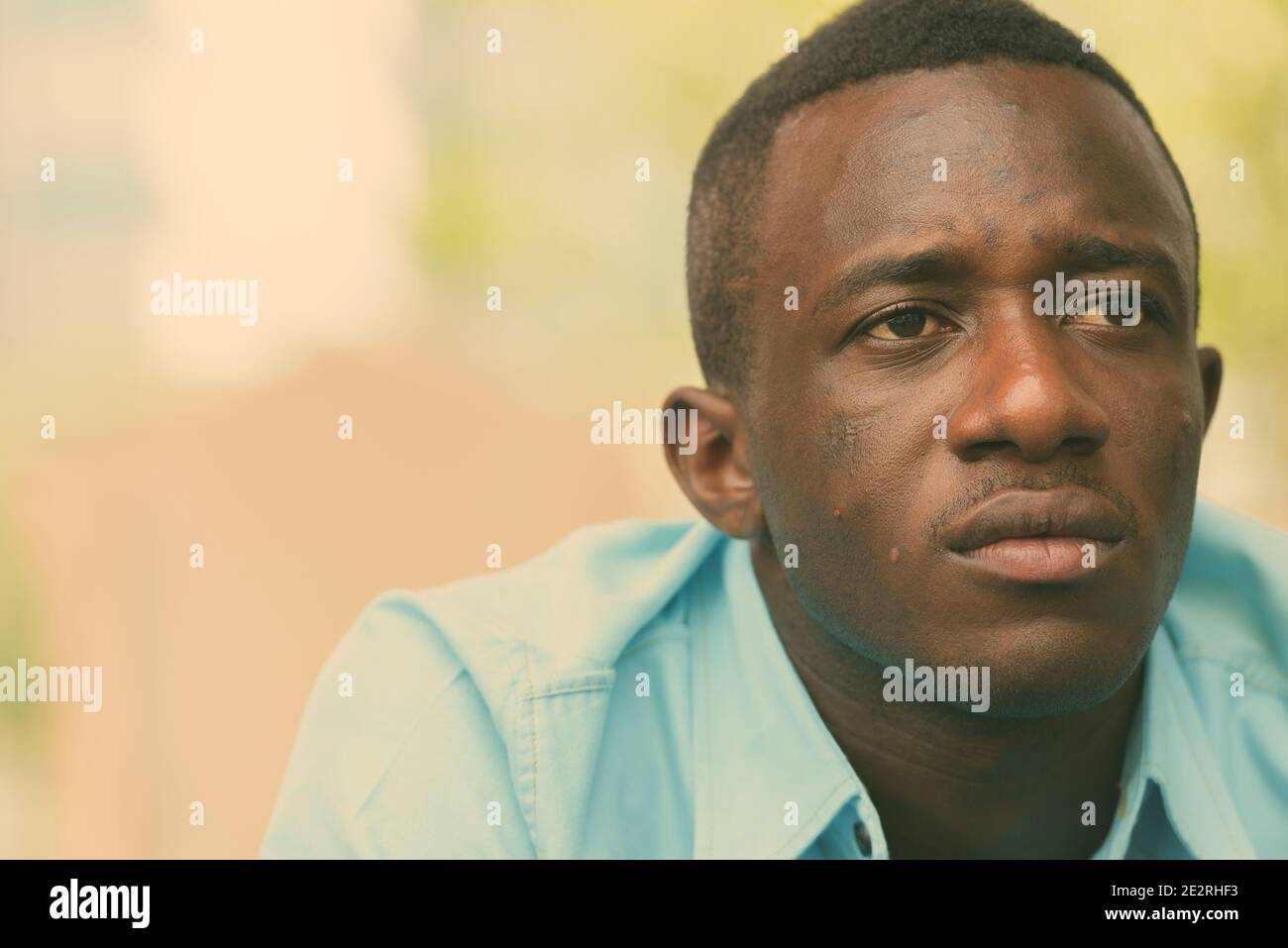 Close up of young black African man sitting while thinking outside coffee shop against view of nature and city Stock Photo