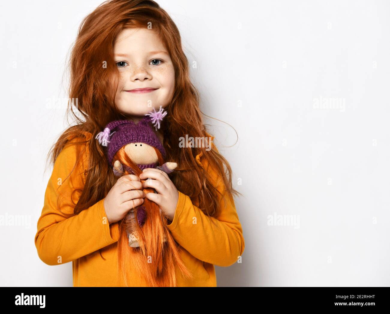 Little five-year-old red-haired kid girl in an orange sweatshirt smiles happily holding small redhair doll in her hands Stock Photo