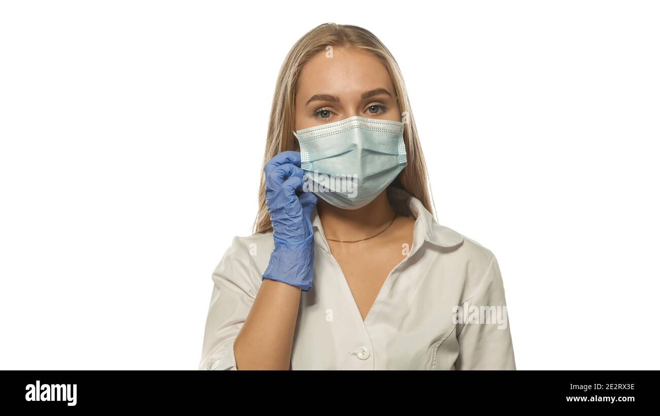 Putting on a face medical mask young blond nurse looking at the camera wearing white medical uniform and disposable blue rubber glows isolated on Stock Photo