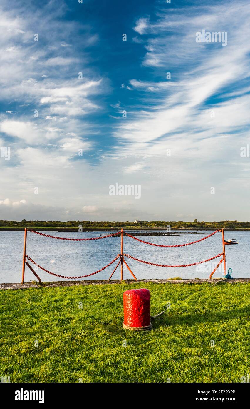A peaceful evening on the quayside at Kinvara Bay, Kinvara, County Galway, Ireland, with red bollard set against green grass and blue sea and sky Stock Photo