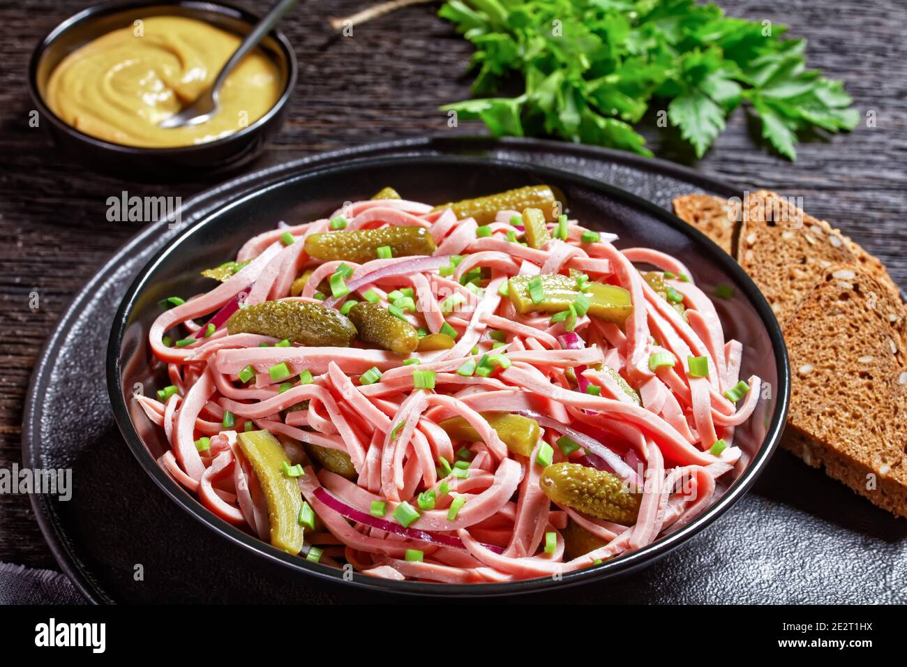 German wurstsalat of cooked german lyoner sausage with pickled cucumbers, red onion slices, vinegar dressing served on a black bowl on a dark wooden b Stock Photo