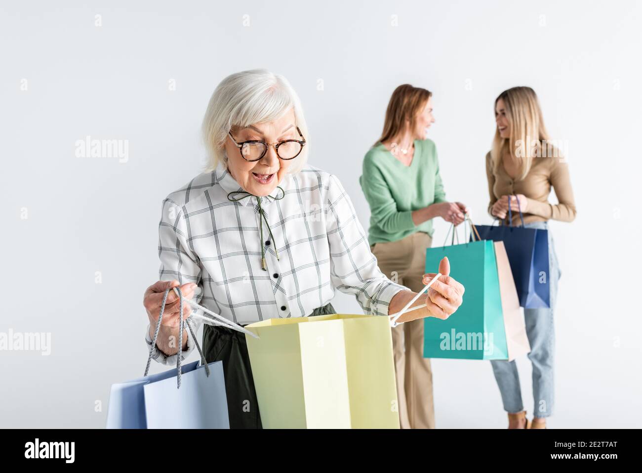 surprised senior woman looking at shopping bag near daughter and granddaughter on blurred background Stock Photo