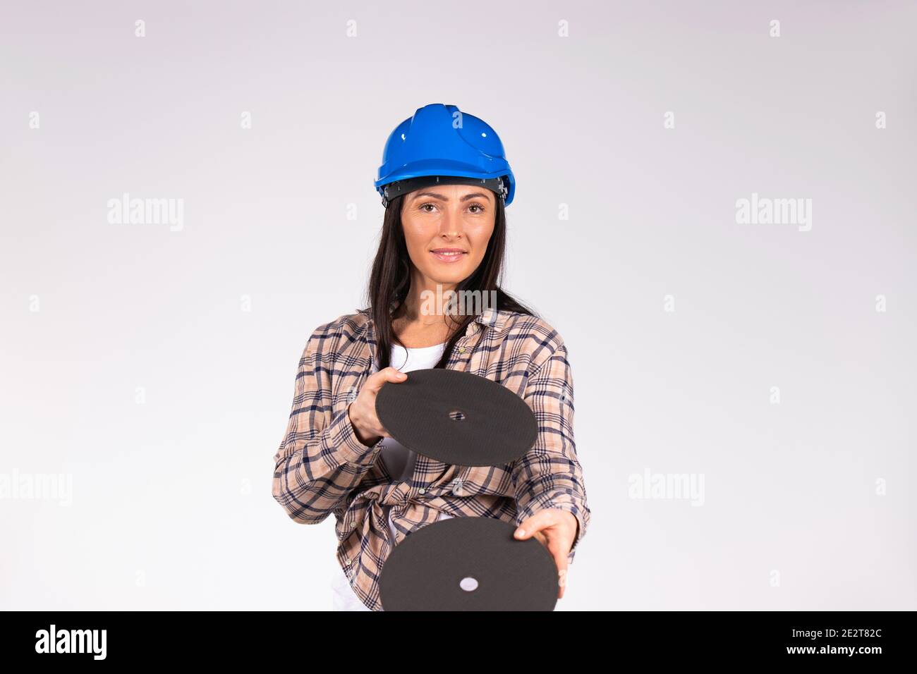 Young brunette in hard hat posing with abrasive discs on a white background. The concept of gender stereotypes. Stock Photo