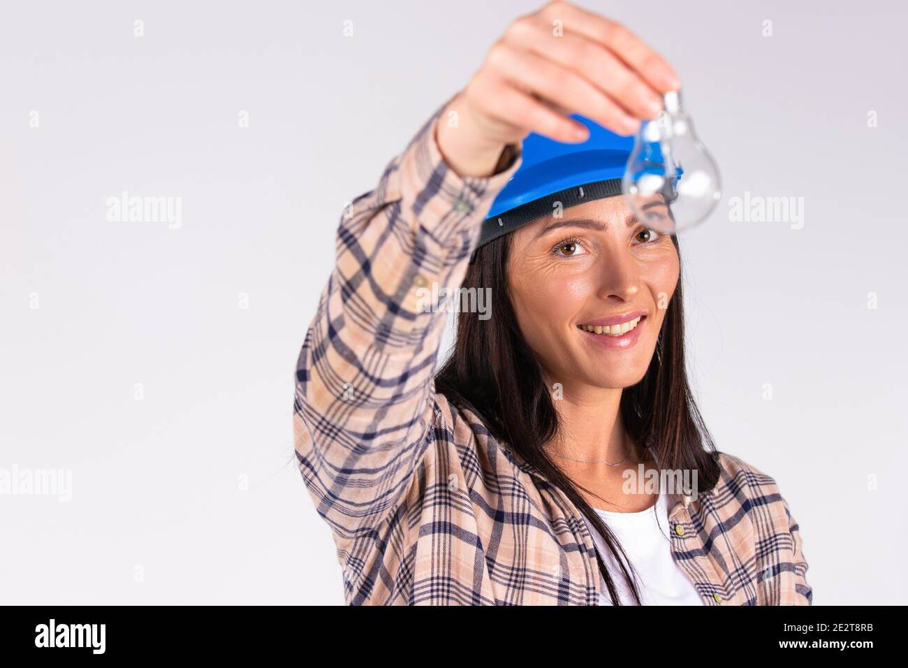 Young brunette in hard hat posing with light bulb and smiling on white background with side space. Stock Photo