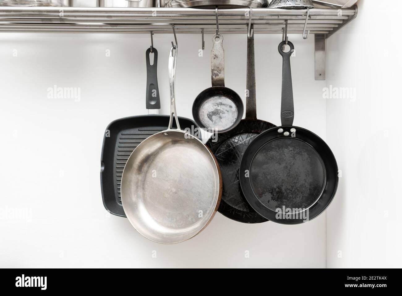 Various pans in different sizes and forms for cooking and frying hanging on metal hooks from shelf in kitchen with white wall in background Stock Photo