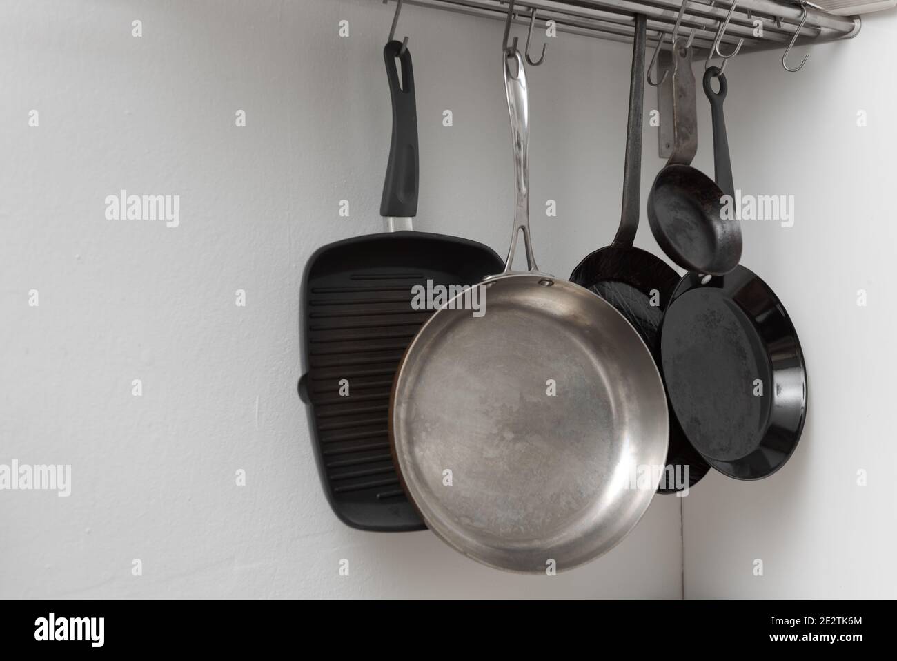 Various pans in different sizes and forms for cooking and frying hanging on metal hooks from shelf in kitchen with white wall in background Stock Photo