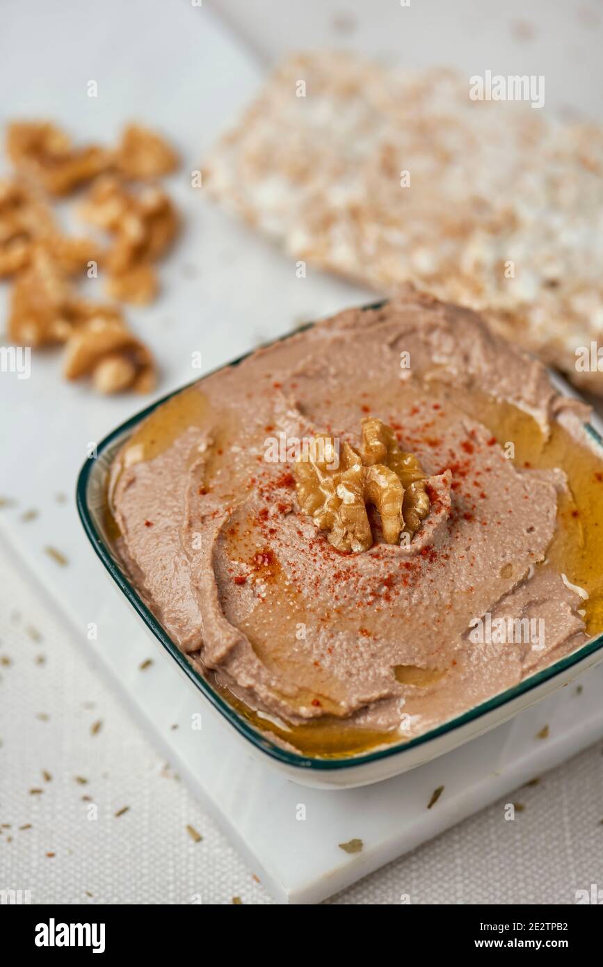 a white ceramic bowl with an appetizing hummus, made with chickpeas, walnuts and rosemary, next to some puffed rice cakes on a white table Stock Photo
