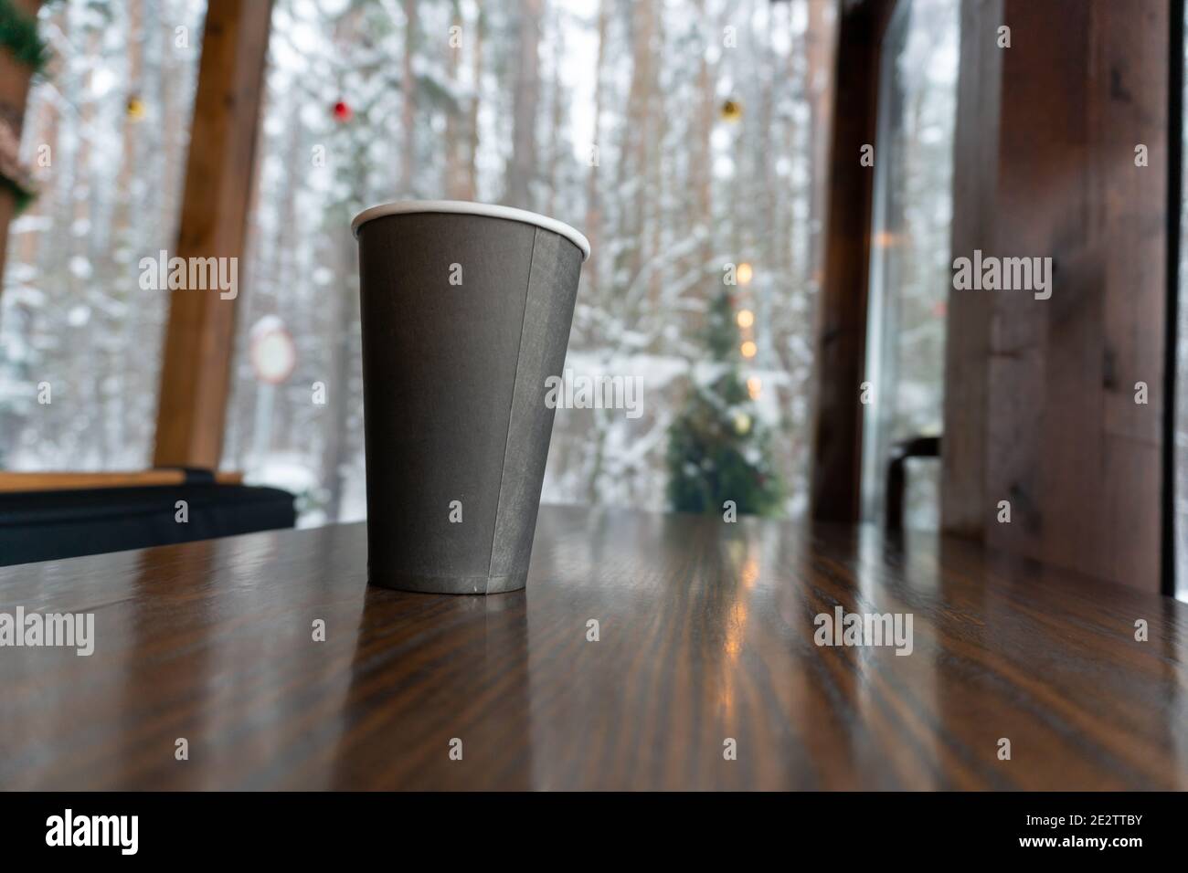 A paper cup of coffee stands on a wooden counter in a pub and a winter forest is visible in the window Stock Photo