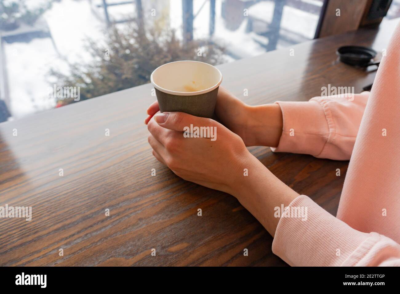 A paper cup of coffee in the hands of a girl in a cafe behind a bar with a view of a winter street Stock Photo
