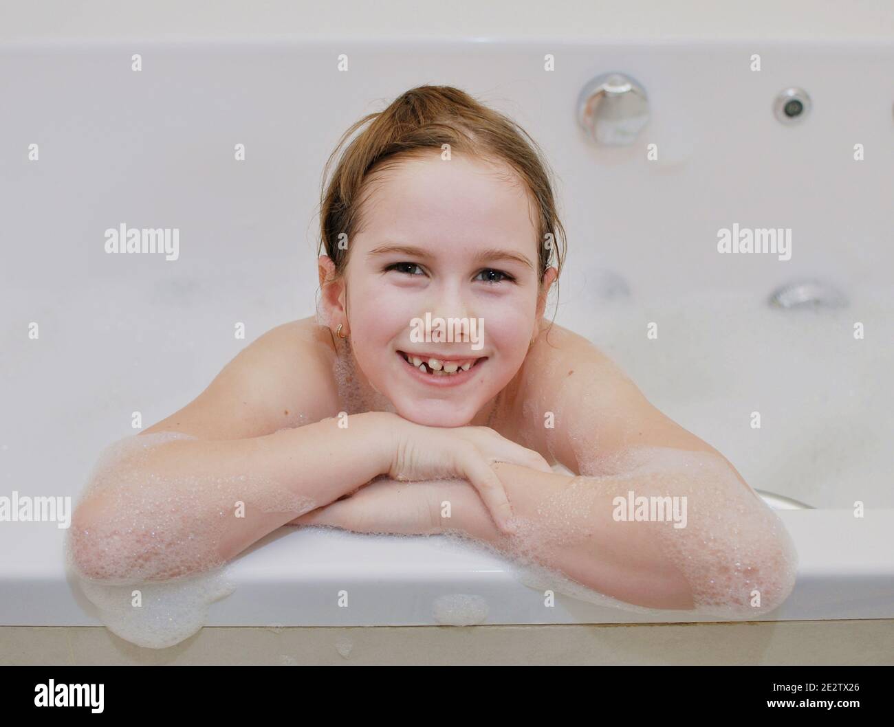 Portrait of an white Caucasian girl (child, kid) smiling with her hands on the edge of the bathtub in the bathroom. Front view Stock Photo