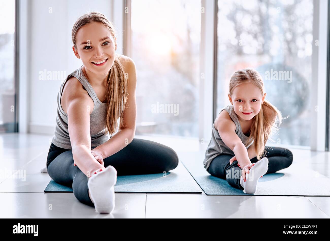 Mom and Daughter are doing yoga. Family in a gym. Little girl with mother in a gray tshirts and black leggings. Girls sitting on a mat and stretching Stock Photo
