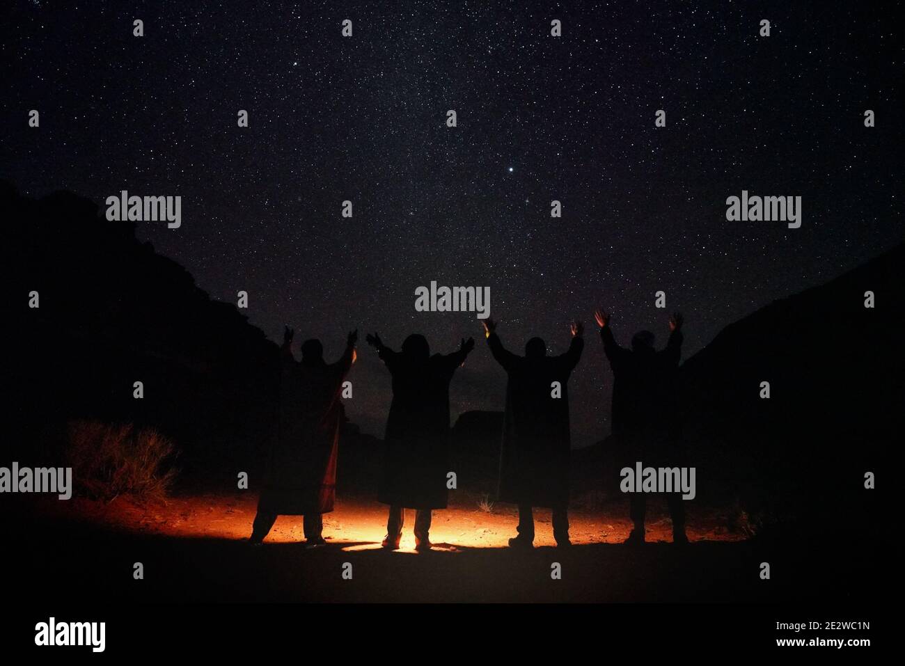 Four persons wearing long warm coats standing in night desert, hands raised to sky with stars, light on the ground, view from behind only silhouettes Stock Photo