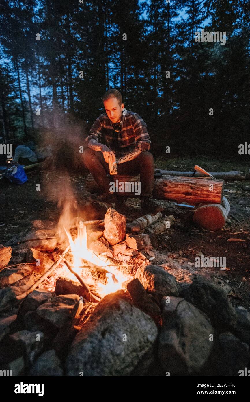 Young Man In Flannel Holding Beer Sits By Campfire At Night In Maine 