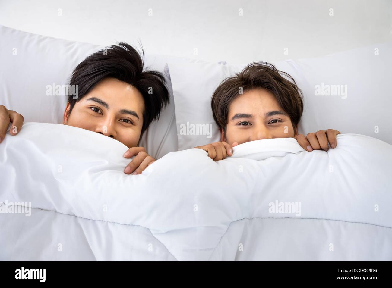 Happy Asian homosexual gay men male couple hiding under blanket in bed together. LGBT and sexual diversity concept. Looking at camera. Stock Photo