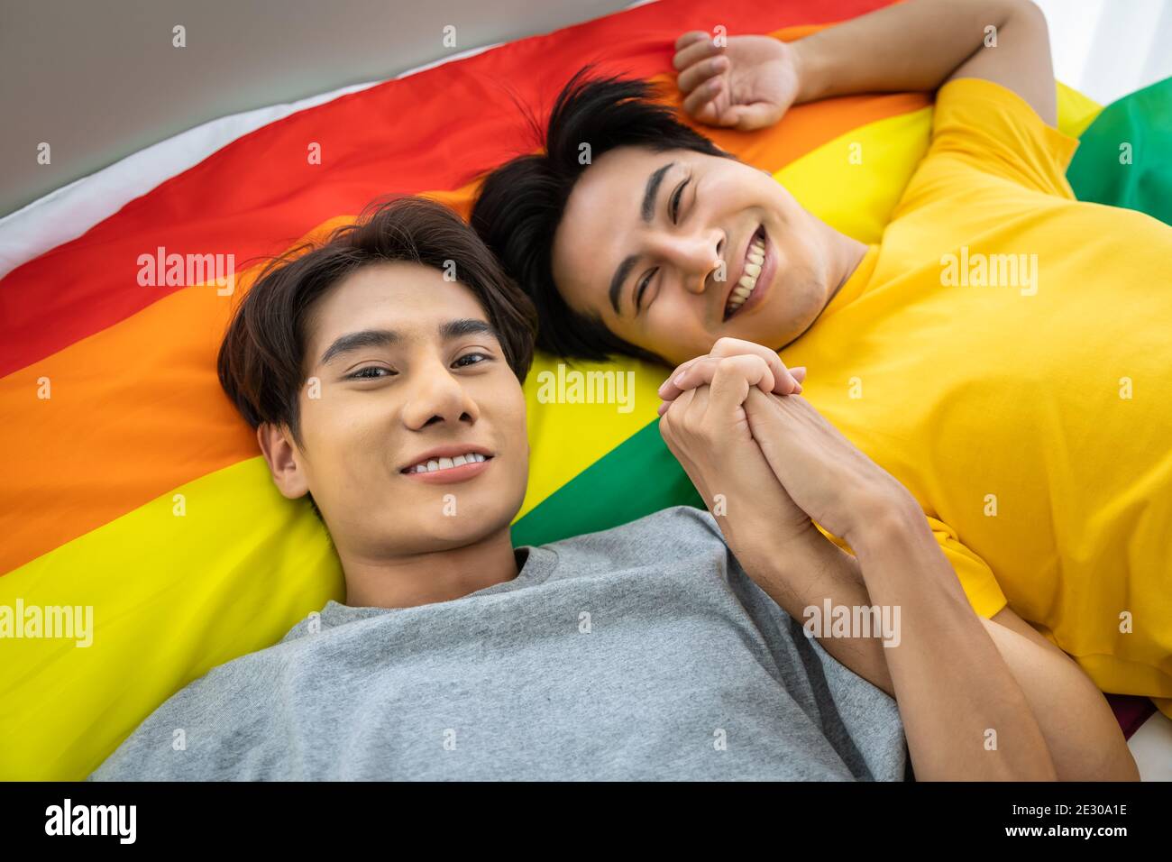Happy Asian homosexual gay couple lying on rainbow pride flag, holding hand. LGBT concept. Looking at camera. Stock Photo