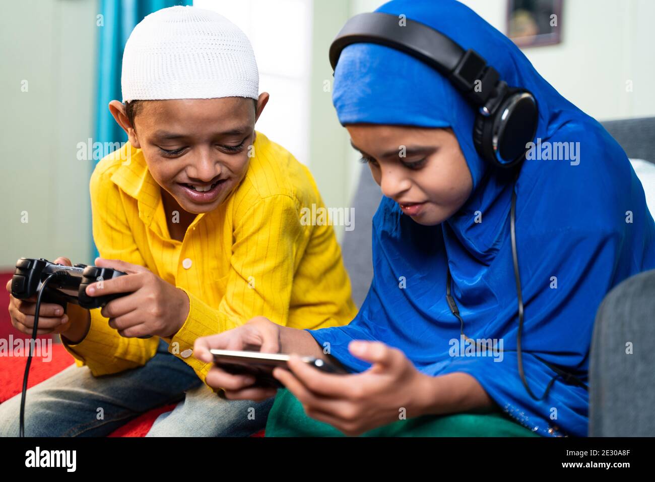 Concept of Kids busy with modern day technology lifestyle and distraction - Two muslim siblings, Brother playing game using joystick and sister playin Stock Photo