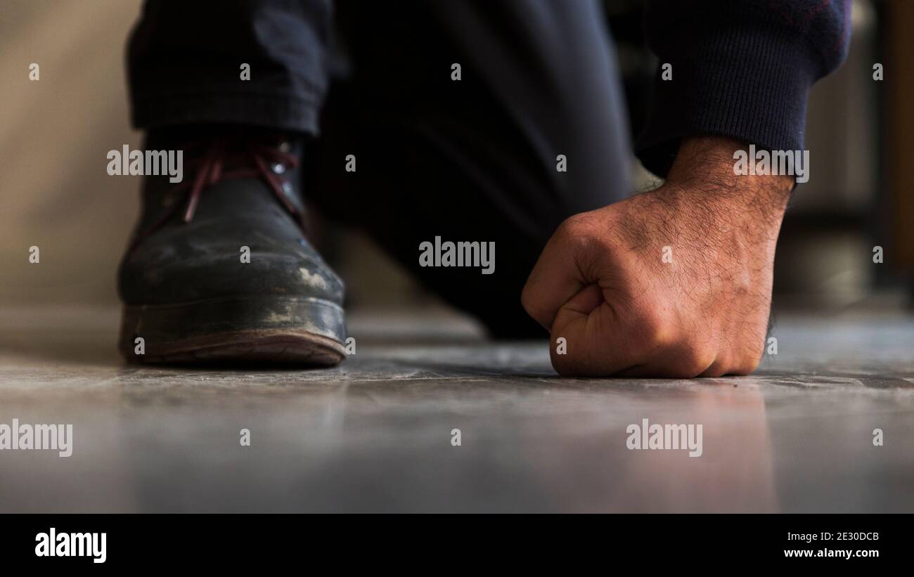 Close up detail of a crouched unrecognizable man's fist and foot with boot, standing on gray, screed floor in front shot. Stock Photo