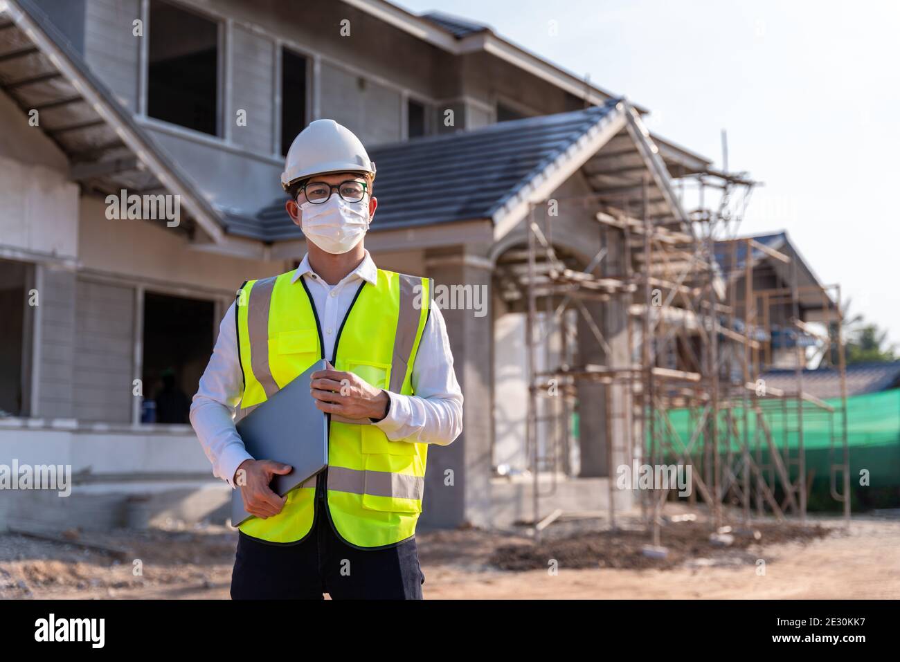 Architect wearing a mask hold a laptop on a building construction site, Homebuilding Ideas and Prevention of Coronavirus Disease. Stock Photo