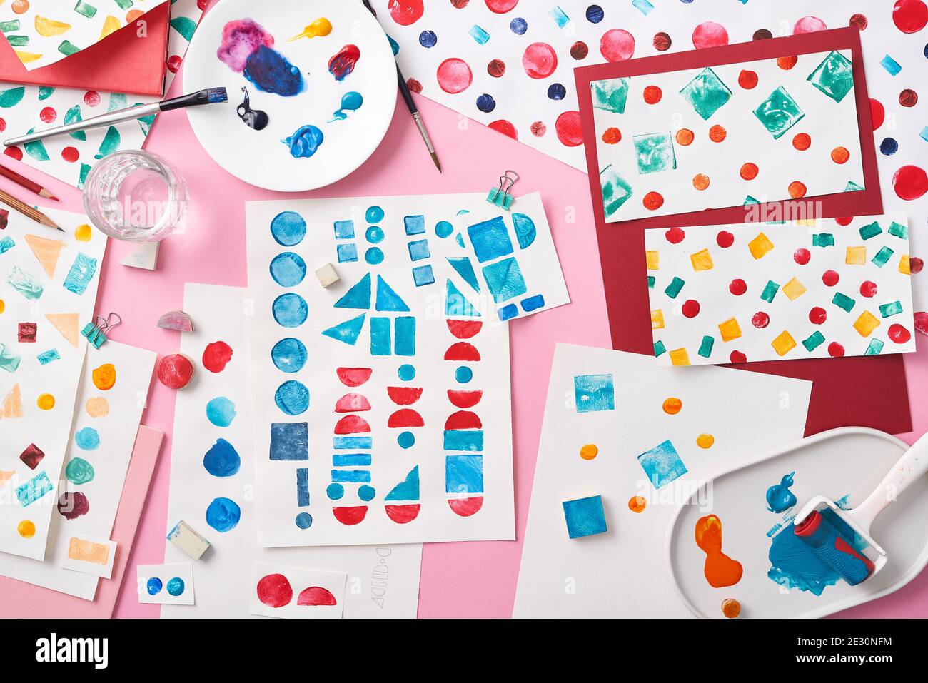 Top view of plates with stains of colorful paint placed on pink background amidst sheets of paper with abstract geometric patterns Stock Photo