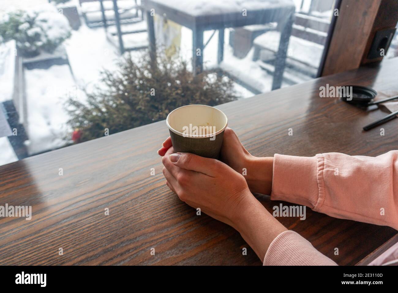 A girl with two hands holds a paper cup with coffee in a cafe overlooking a winter street Stock Photo