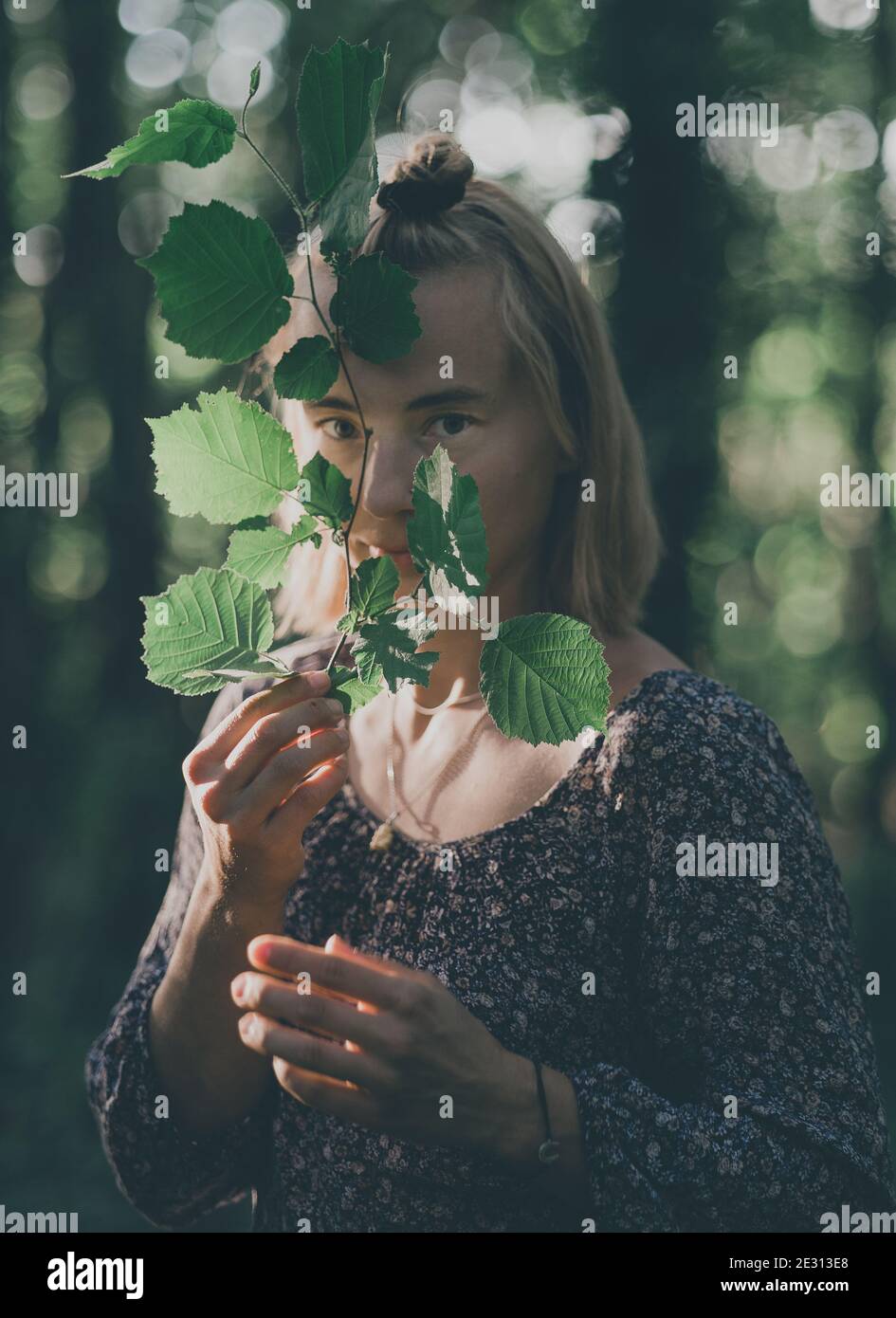 A woman peers through a leafy branch standing in a forest, backlit by the setting sun during golden hour Stock Photo