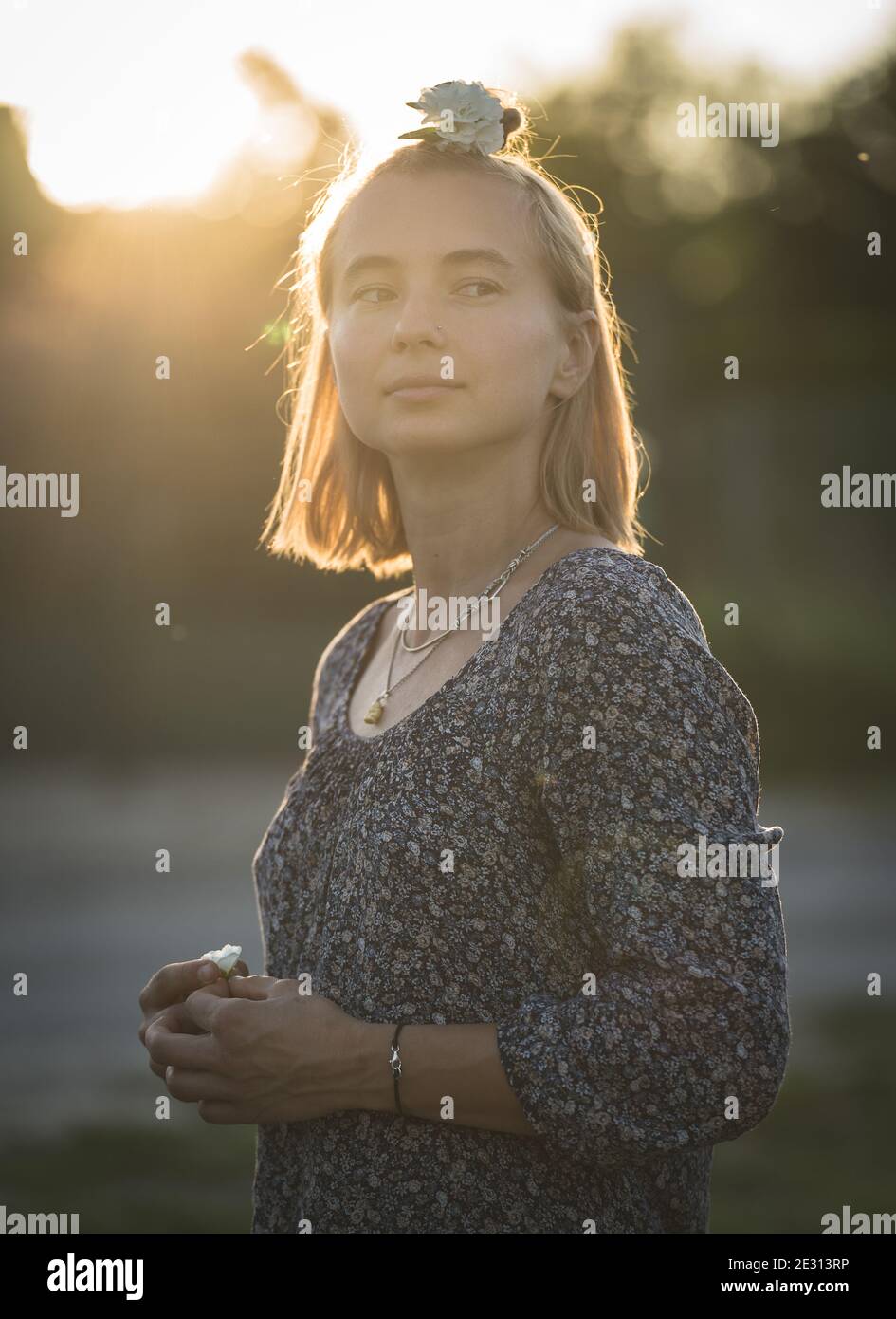 A woman with blonde hair wears a white flower in her hair, backlit by ...