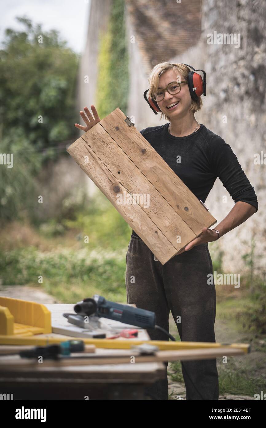 A woman wearing ear defenders proudly holding a piece of wooden furniture that she is building. Stock Photo