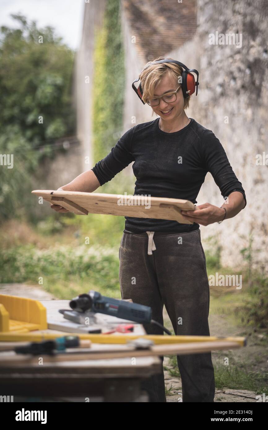 A woman wearing ear defenders proudly holding a piece of wooden furniture that she is building. Stock Photo