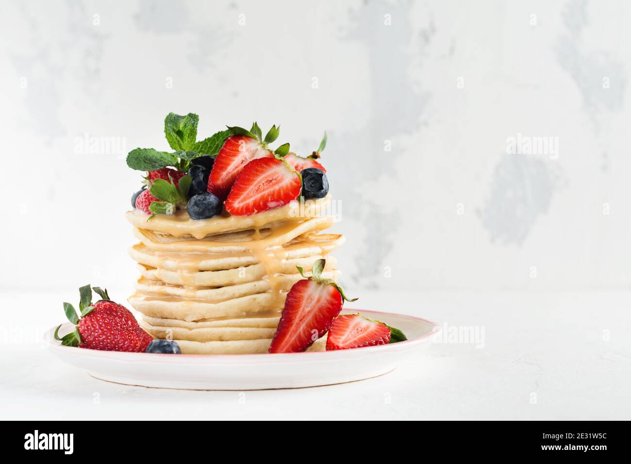 Stack of homemade little pancakes with honey, fresh strawberries and sauce on light wooden background. Selective focus. Stock Photo