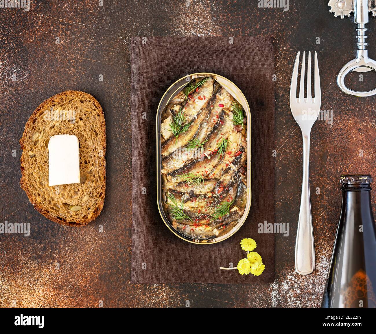 Open tin of Baltic sprats in oil with spices, a slace of bread with butter, a fork and bottle of dark beer on an old brown background. Fish appetizer Stock Photo